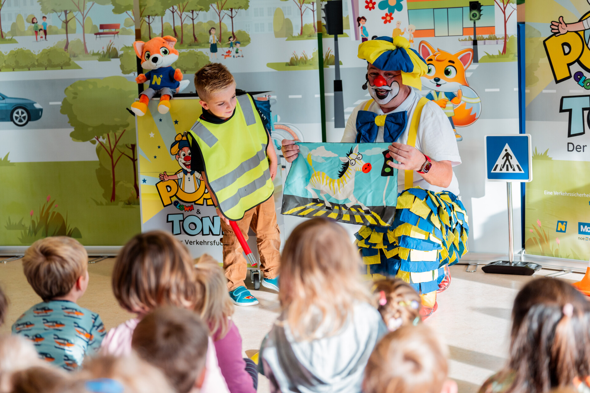 Verkehrsclown Poppo zeigt ein Zebra auf dem Zebrastreifen, Kind daneben schaut staunend hin und Kinder vor Bühne sind gespannt