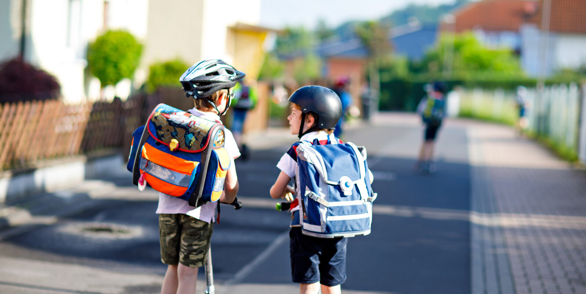 Two school kid boys in safety helmet riding with scooter in the city with backpack on sunny day. Happy children in colorful clothes biking on way to school.