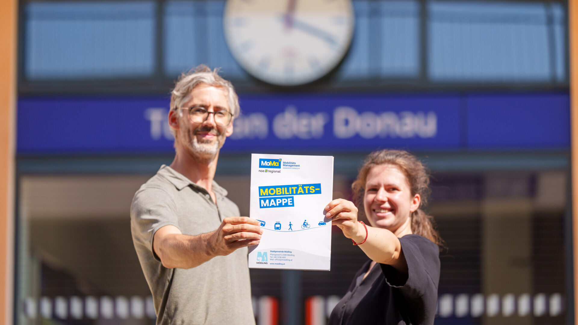 Zwei Personen halte Mappe in die Kamera, im Hintergrund ein Bahnhof