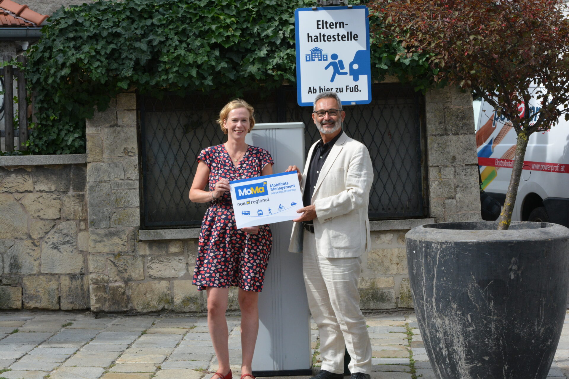 Verkehrsberaterin und Vizebürgermeister stehen mit Fototafel vor einem Elternhaltestellenschild