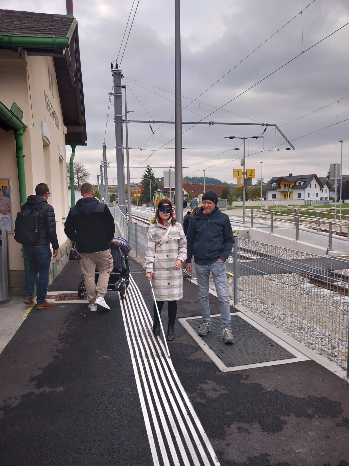 Personen probieren sich mit Blindenstock und Kinderwagen den Barrieren eines Bahnhofs zu stellen.